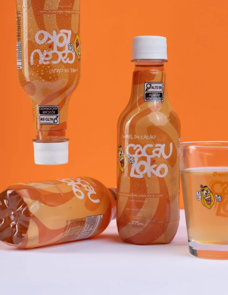 Garrafas de Cacau Loko em pé e deitadas ao lado de um copo da bebida, sobre fundo laranja