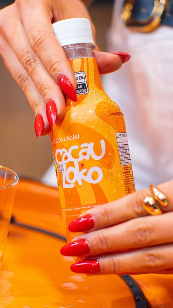 Mãos de unhas vermelhas segurando a garrafa de Cacau Loko ao lado de um copo